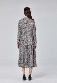 Femme se tenant de dos, portant un chemisier à manches longues blanc à fleurs noires et une jupe midi assortie avec des bottines noires.