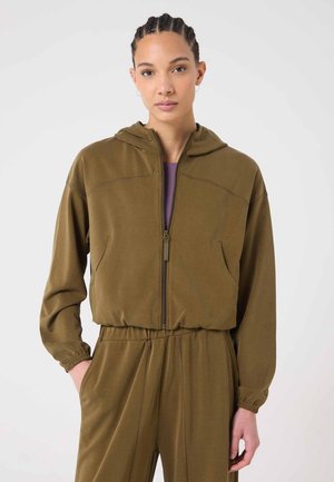 Femme aux cheveux tressés portant un sweat à capuche olive à fermeture éclair et un pantalon assorti, les mains dans les poches, fond neutre.