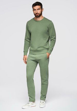 Mann mit Bart, der einen passenden olivgrünen Pullover und Jogginghosen sowie weiße Sneakers trägt, steht mit einer Hand in der Tasche.