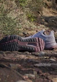 Scarpa da trail bianca e viola distesa su terra rocciosa, che mostra una suola testurizzata con il testo "TRAXION", circondata da vegetazione secca.