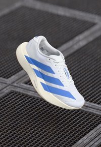 Pantofii albi sport, cu trei dungi albastre, superior din plasă texturată și talpă de culoare crem. Design ușor, potrivit pentru alergare.