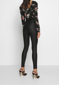 Leggings en simili cuir noir au design moulant, associés à un blouse noir à fleurs avec des manches volantées et un dos ouvert. Des talons hauts sont portés.