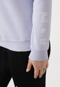 Helllila Sweatshirt mit gerippten Bündchen und Saum. Auf dem Ärmel befindet sich der Aufdruck "WORLD SERIES OF FITNESS RACING" und "NEVER STOP COMPETING."