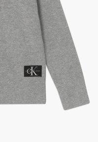 Šedý žebrovaný svetr s černou nášivkou loga Calvin Klein Jeans na boku. Materiál je měkký a pletený s přiléhavým střihem.