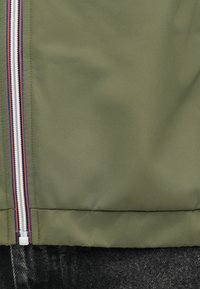 Veste vert olive en tissu lisse avec une fermeture éclair blanche comportant des accents rouges et bleus. Inclut une couture horizontale en bas.
