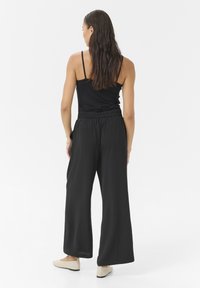 Schwarze weit geschnittene Hosen aus leichtem Material mit elastischem Bund und seitlichen Taschen, kombiniert mit einem enganliegenden schwarzen Tanktop und flachen Schuhen.
