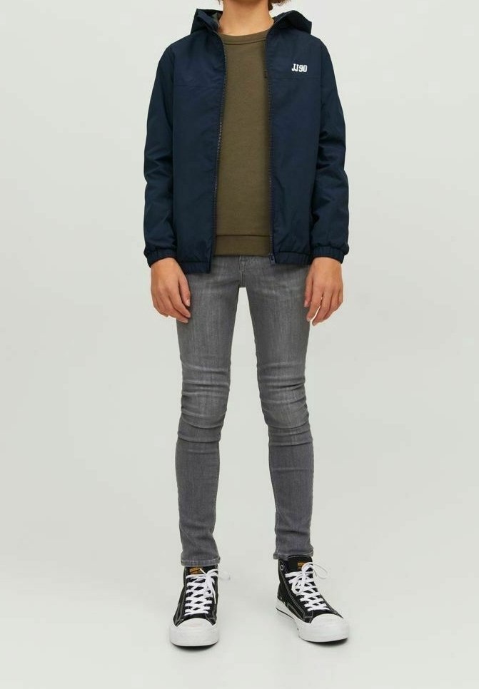Enfant portant une veste bleu marine, une chemise vert olive, un jean skinny gris et des baskets montantes noires avec des lacets blancs, debout devant un fond uni.