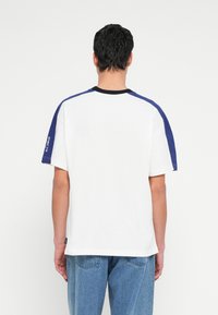 Tricou alb din bumbac cu mâneci bleumarin, guler negru și un detaliu vizibil cu logo pe mâneca stângă. Croială lejeră, tip cutie.