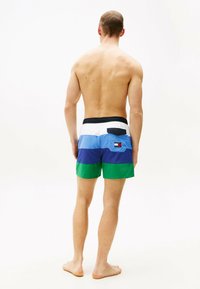 Shorts de bain colorés avec un design patchwork, présentant des sections bleu, vert et blanc. Ceinture élastique noire, poche arrière avec un logo.