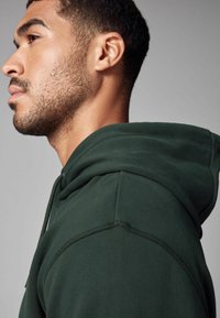 Green hoodie featuring a textured fabric, a lined hood, and visible stitching details. Close-up view focuses on the shoulder and neckline.