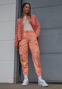 Korallfärgad denimskjorta, beige rynkad topp, orange cargobyxor med meshdetaljer och sneakers med pastellfärger samt vita strumpor.