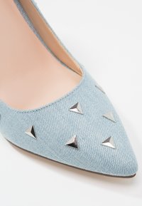 Escarpin en denim bleu clair à bout pointu, agrémenté de clous triangulaires en argent sur le dessus pour plus de détail et de texture.