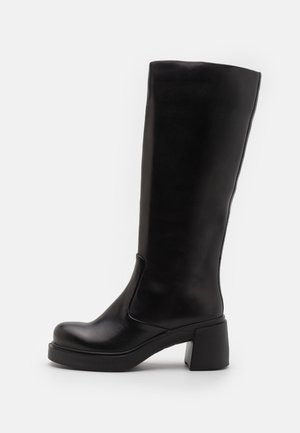 Bota preta até ao joelho feita de couro liso, com um salto robusto e biqueira arredondada. Design simples com um elástico lateral para facilitar o calçar.