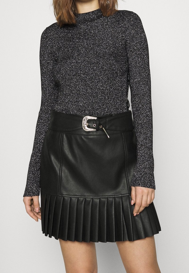 Jupe plissée en cuir noir avec ceinture à boucle argentée, associée à un haut en maille métallisée foncée à manches longues et col montant.