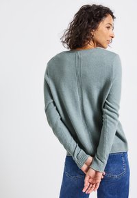 Pull vert clair en matériau doux, avec un col rond côtelé, des coutures latérales et des poignets et un ourlet côtelés. Porté avec un jean en denim bleu.
