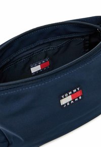 Pochette blu navy con chiusura a zip, caratterizzata da una texture tessile e un'etichetta "TOMMY JEANS" in rosso, bianco e blu all'interno.