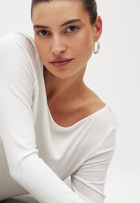White ribbed long-sleeve top with a wide neckline, paired with hoop earrings. Smooth texture, fitted design, minimalistic style.