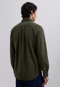 Man wearing a dark green checked long-sleeve shirt and blue jeans, shown from the back against a plain light background.