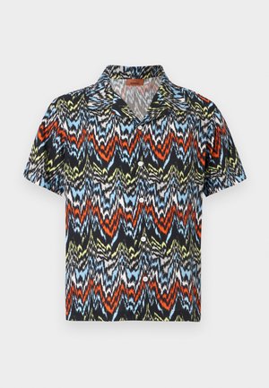 Mand, der står op ad en ensfarvet væg iført en flerfarvet t-shirt med zigzag-mønster, opkørte mørke jeans og brune sandaler.