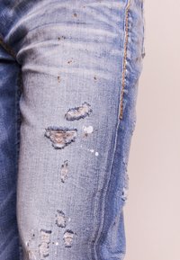 Světlé modré džíny s opotřebovaným vzhledem, zahrnující roztržené okraje, stříkance barvy a vybledlou texturu. Kontrastní šití podél švů.