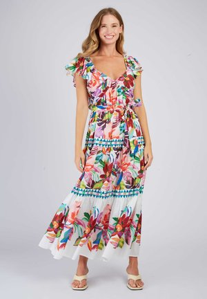 Smiling woman wearing a colorful floral maxi dress with ruffled sleeves and a waist tie, standing against a plain light background.