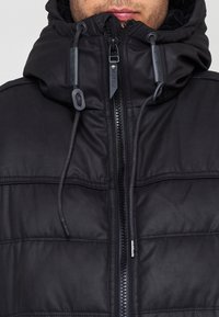 Giubbotto imbottito nero con colletto alto, chiusura con zip e dettagli con cordini. Presenta una texture liscia e lucida e dettagli di cuciture orizzontali.
