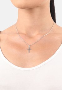 Nahaufnahme einer Person, die eine silberne Kreuzanhänger-Halskette auf einer dünnen Kette über einem weißen Rundhalsausschnitt-Shirt trägt.