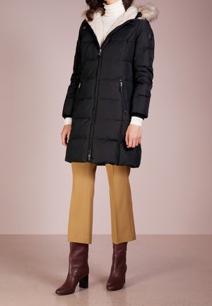 Zwarte gewatteerde parka met een met nepbont gevoerde capuchon, rits aan de voorkant, twee ritszakken en geribbelde manchetten. Combinatie met beige broek en bruin schoenen.