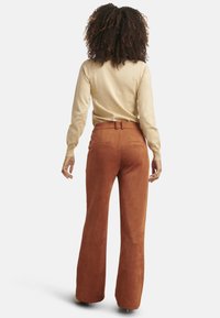 Brown suede flared pants with a high waistband and rear pockets, paired with a beige long-sleeve top, seen from the back.