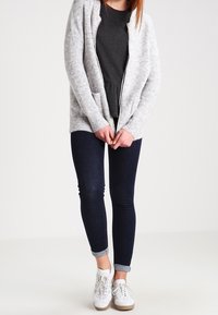 Hellgraue gestrickte Strickjacke mit Taschen, getragen über einem dunklen Oberteil, kombiniert mit dunklen Skinny-Jeans und weißen Sneakern. Einfaches, strukturiertes Design.