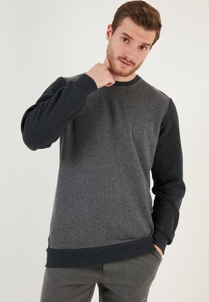 Felpa grigia con un motivo a spina di pesce testurizzato sul davanti e maniche scure uniformi. Collo e polsini a coste. Tessuto morbido, vestibilità casual.