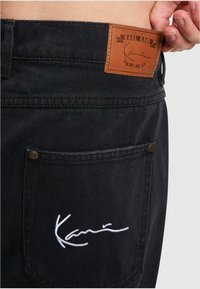 Los pantalones de mezclilla negros presentan una etiqueta de cuero con la marca y una firma bordada en blanco en el bolsillo trasero, destacando la textura y el detalle.