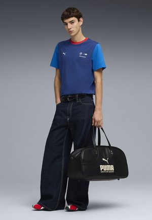 Joven con camiseta azul de BMW Motorsport, vaqueros anchos oscuros, zapatos rojos y blancos, sosteniendo una bolsa deportiva negra grande de Puma.