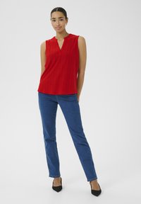 Rotes ärmelloses Oberteil mit V-Ausschnitt, das mit Lochmustern verziert ist. Kombiniert mit blauen geraden Jeans und schwarzen spitzen Absatzschuhen.