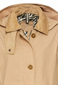 Beige Mantel mit einem Cordkragen, zwei Knöpfen und einem Reißverschluss; hat ein Innenfutter mit Zebramuster. Glattes Gewebe mit leichter Textur.