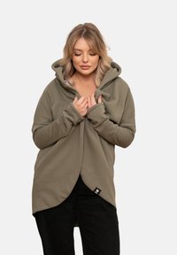 Oliwkowa bluza z kapturem wykonana z miękkiego materiału, z dużym kapturem, długimi rękawami i skośnym zapięciem z przodu oraz detalem metki po boku.