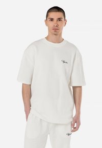 White ribbed cotton t-shirt with short sleeves, relaxed fit, and embroidered logo on the left chest. Textured surface, round neckline.