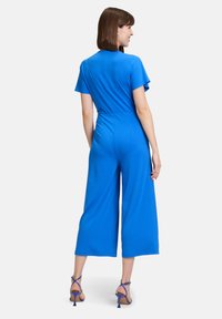 Giacca blu brillante con maniche corte, vita aderente, pantaloni larghi, realizzata in tessuto morbido. Abbinata a tacchi blu con cinturini.