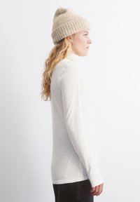 Balta garo piedurkņu turtlenecks, kas izgatavots no mīksta, viegla auduma. Modelis valkā bēša adītu cepuri ar ribotu maliņu. Sānu profils.