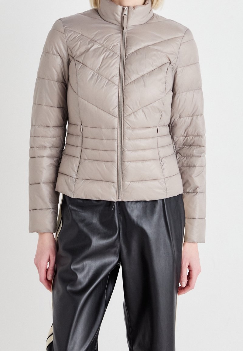 Puff jacket in light beige, quilted with chevron pattern. Features a stand-up collar and full front zipper. Paired with black faux leather pants.