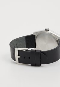 Correa de reloj de cuero negro con herrajes metálicos plateados. Cuenta con una tapa trasera de acero inoxidables y detalles del logo en el lado interior de la correa.
