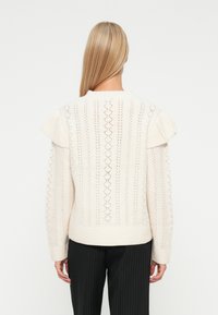 Kobieta z długimi blond włosami, ubrana w beżowy sweter dzianinowy z falbaniastymi detalami na ramionach i czarne spodnie w prążki, zwrócona tyłem.