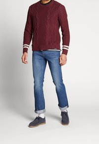 Burgunderfärgad stickad tröja med kabelmönster och vita ränder på ärmarna, kombinerad med ljusblå uppvikta jeans och mörkgrå mockaskor.