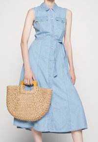 Jasnoniebieska dżinsowa sukienka bez rękawów z kołnierzykiem i zapięciem na guziki z przodu, wysmuklona w talii, z dwiema kieszeniami na piersi. W dłoni trzymana jest pleciona torba w kształcie półksiężyca.