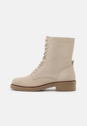 Bottine en daim beige avec fermeture à lacets à l'avant, semelle marron avec petit talon bloc, et languette arrière, sur un fond clair uni.