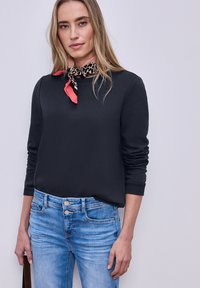 Femme portant un haut noir à manches longues, un jean bleu clair avec double boutons, et un foulard rouge et noir à motifs autour du cou, debout contre un mur uni.