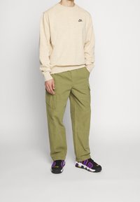 Sweat à col rond en coton beige avec logo noir ; pantalon cargo vert olive avec poches latérales ; baskets noires et violettes.