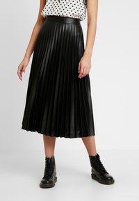 Gonna midi plissettata nera in ecopelle con una texture liscia e lucida. Indossata con una blusa a pois e stivali neri con lacci.