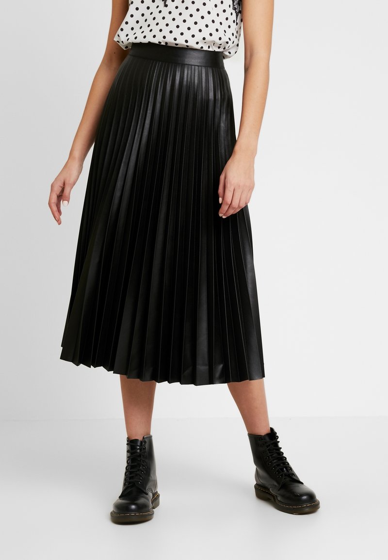 Gonna midi plissettata nera in ecopelle con una texture liscia e lucida. Indossata con una blusa a pois e stivali neri con lacci.