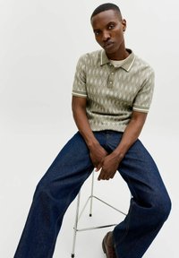 Junger Mann, der ein gemustertes kurzärmeliges Poloshirt und weit geschnittene Jeans trägt und auf einem Metallhocker vor einem schlichten Hintergrund sitzt.